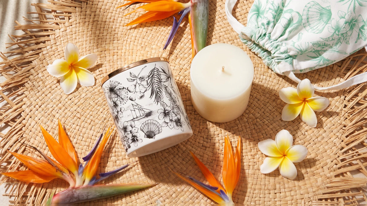 Told London Island Breeze, Seychelles candle in white fine porcelain with hand-drawn black tropical motifs and gold lid, beside its wax refill and shell-print cotton pouch, with frangipani and birds of paradise on woven raffia