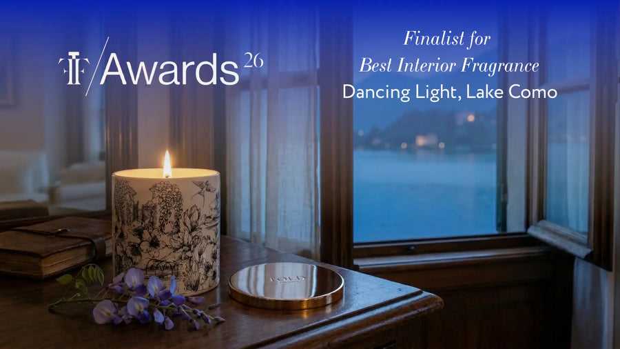 Candela Dancing Light, Lake Como accesa in porcellana bianca con motivi neri disegnati a mano, coperchio dorato e glicine, su una scrivania alla finestra sul Lago di Como al crepuscolo, con il marchio di finalista dei Fragrance Foundation Awards 2026
