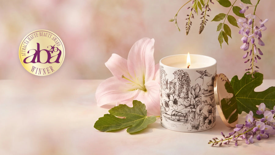 Candela Dancing Light, Lake Como accesa, in porcellana fine bianca con motivi neri disegnati a mano e coperchio dorato, con un giglio rosa, foglie di fico e glicine, e a sinistra il medaglione dorato di vincitore degli Attracta Beauty Awards 2026