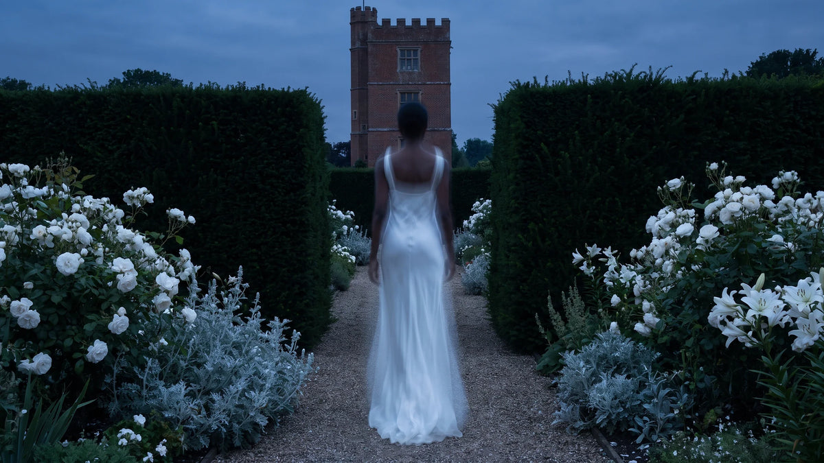 Una figura in abito bianco velato, di spalle, cammina tra rose bianche e siepi di tasso verso una torre di mattoni.