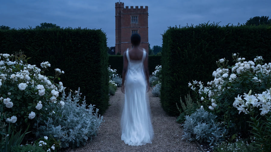 Una figura in abito bianco velato, di spalle, cammina tra rose bianche e siepi di tasso verso una torre di mattoni.