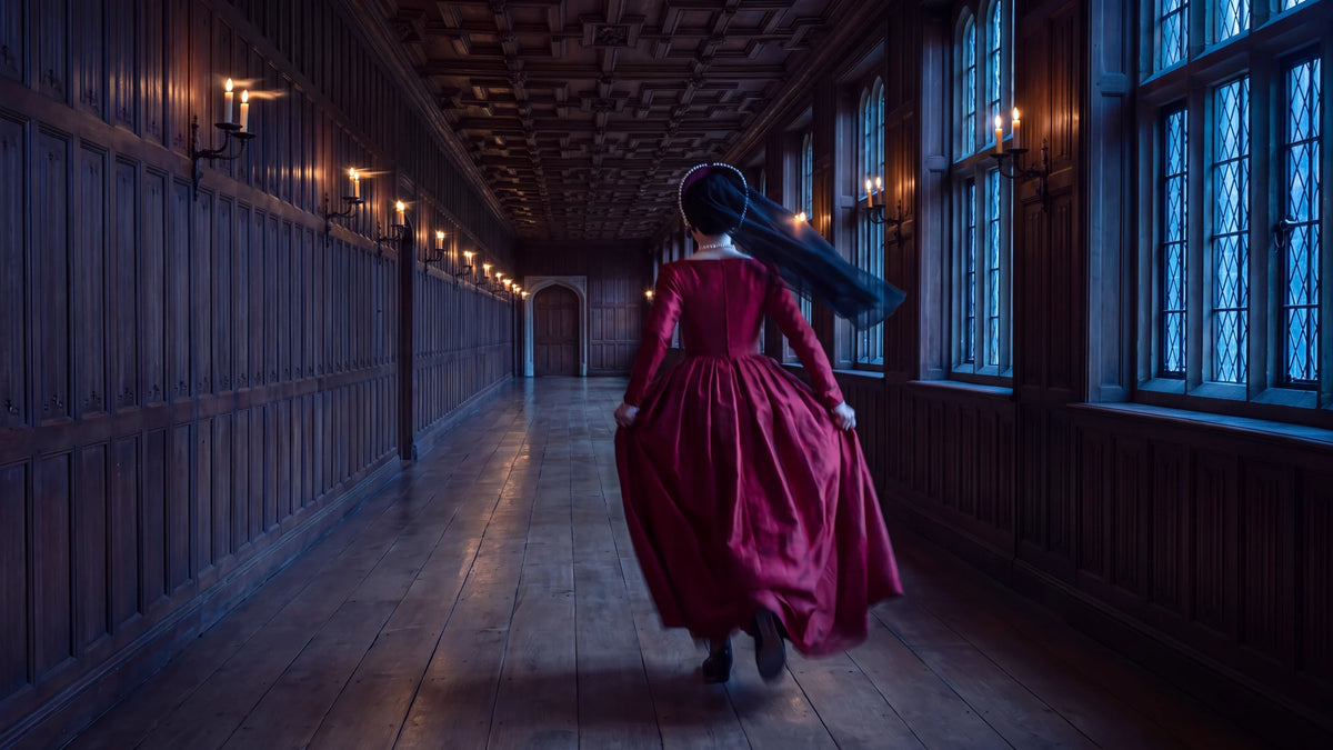 Nainen karmiininpunaisessa Tudor-ajan puvussa ja ranskalaisessa päähineessä takaapäin kuvattuna juoksemassa pitkää paneloitua galleriaa kohti suljettua ovea, kynttilänliekit taipuvat sivulle.