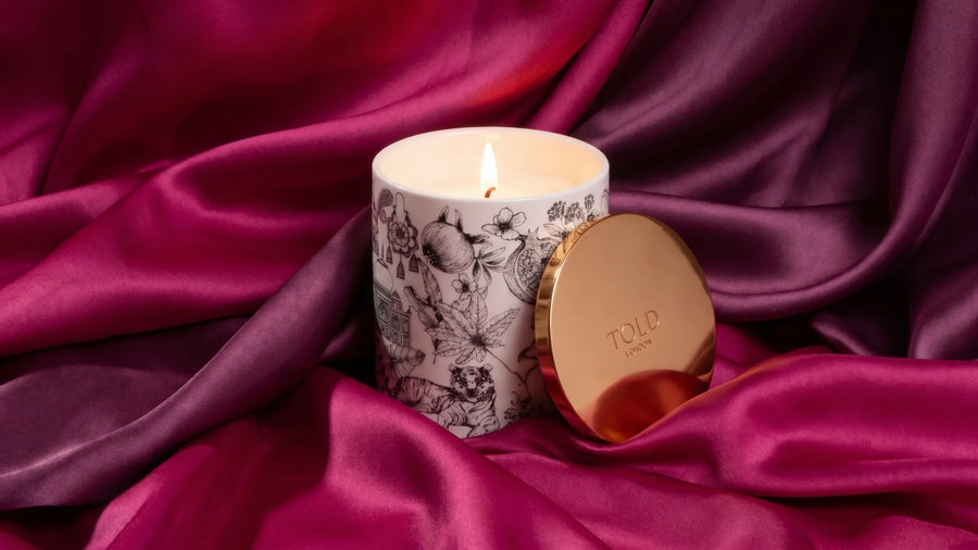 Candela Told London in porcellana fine con motivi illustrati neri disegnati a mano, accesa, il coperchio dorato appoggiato al vaso, tra pieghe profonde di seta magenta e prugna