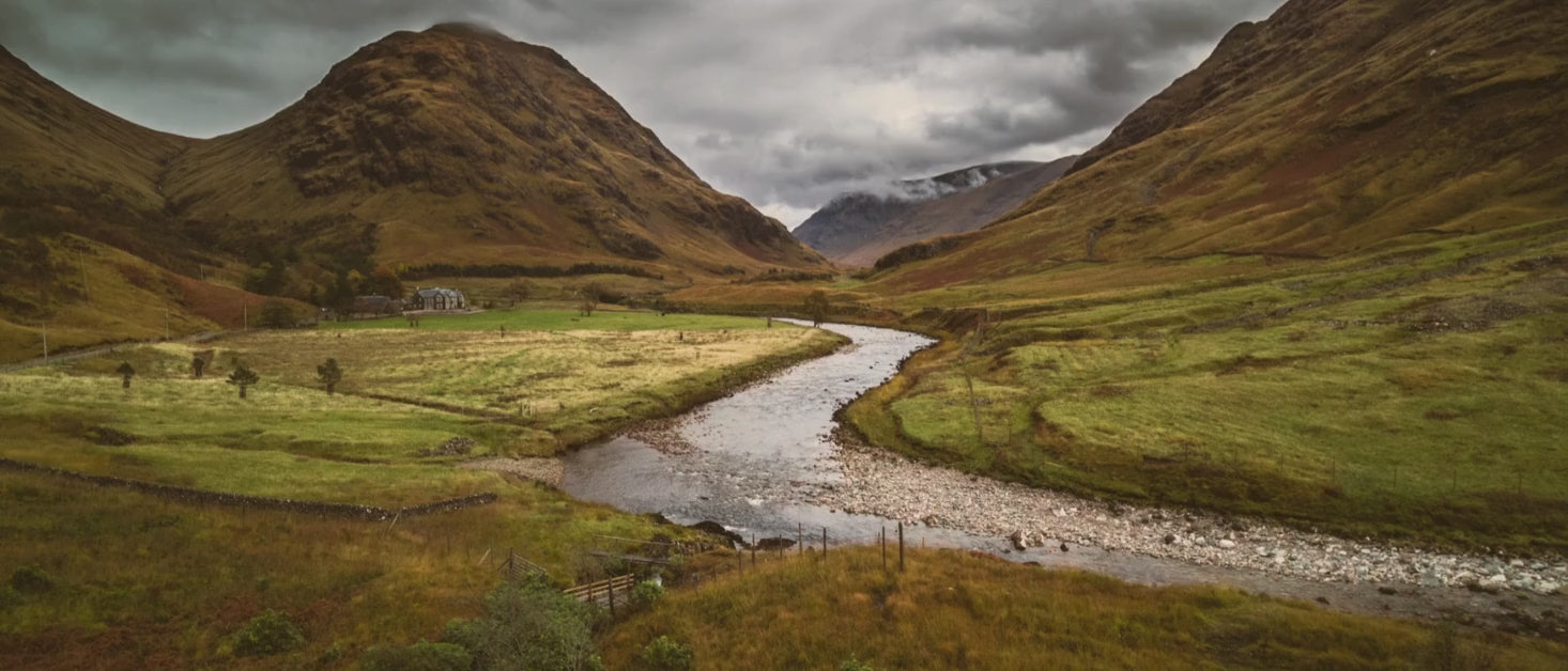 Camera shoots overhead of a scottish valley with low clouds and a house in the distance