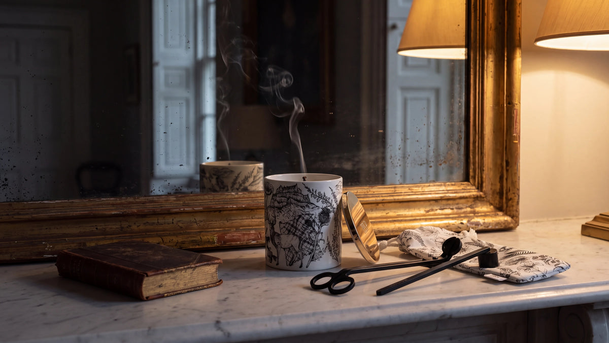 A Told London candle on a marble mantelpiece beneath a gilt mirror, just snuffed with a thread of smoke rising, its gold lid alongside, the black wick trimmer and snuffer and their printed cotton pouch beside an old leather-bound book.