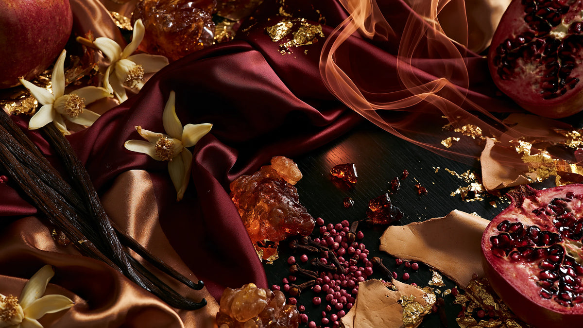 Oxblood and bronze silk scattered with amber resin, split pomegranates, vanilla pods, pink peppercorns, cloves, pale blossoms and gold leaf, with a curl of smoke.