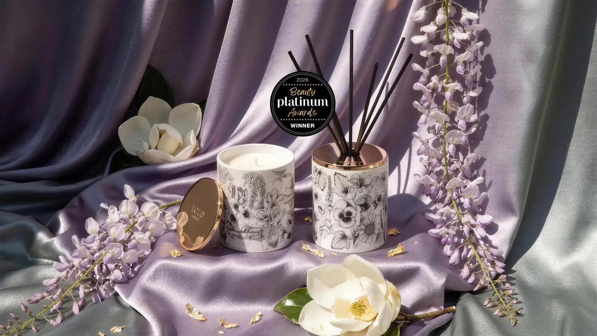 A Told London illustrated porcelain candle and reed diffuser side by side on lilac satin, with trailing wisteria, magnolia flowers and the 2026 Platinum Beauty Awards winner badge above them.