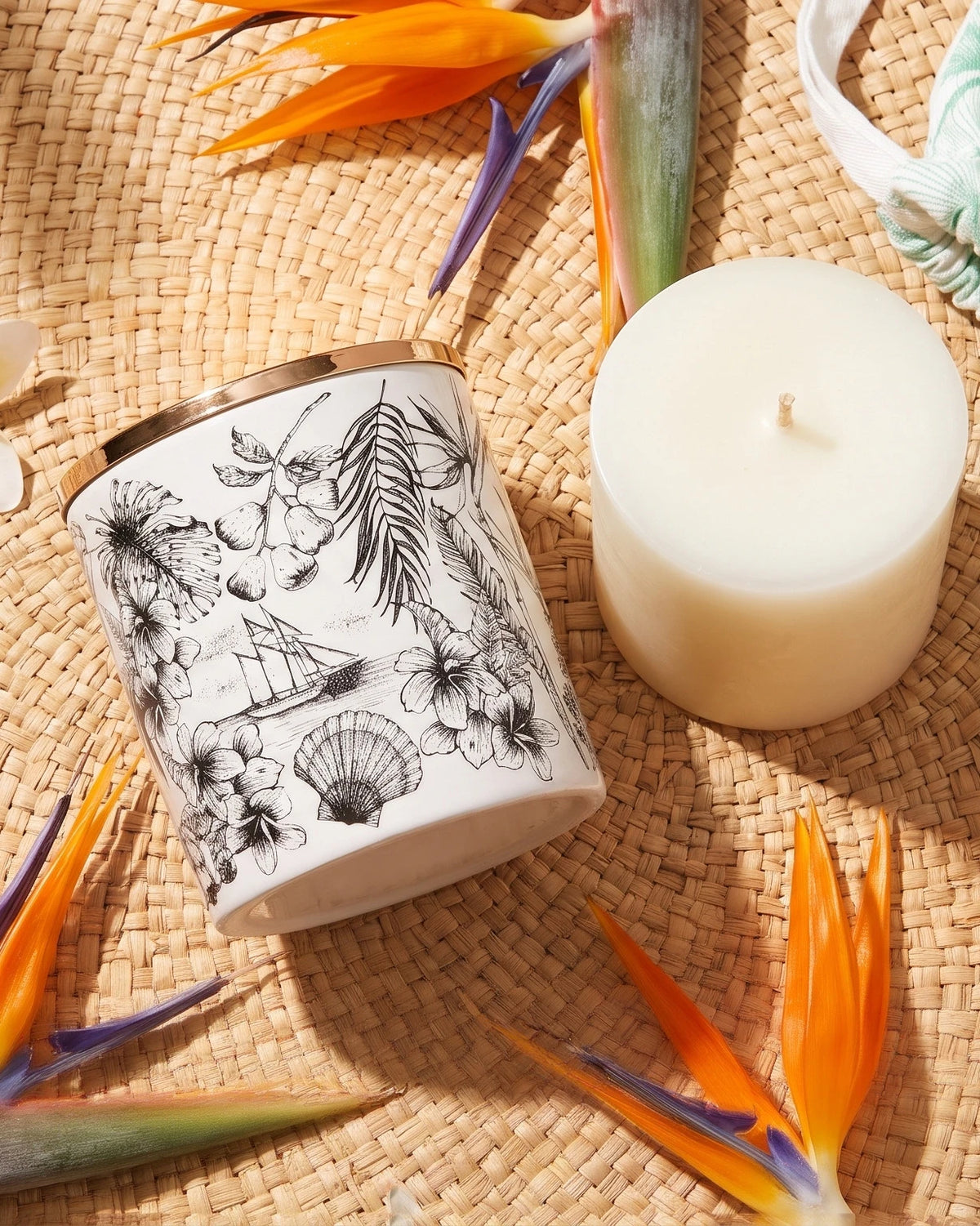 Un vaso in porcellana Island Breeze accanto alla sua ricarica per candela su una stuoia di paglia intrecciata, con frangipane e fiori di strelitzia.