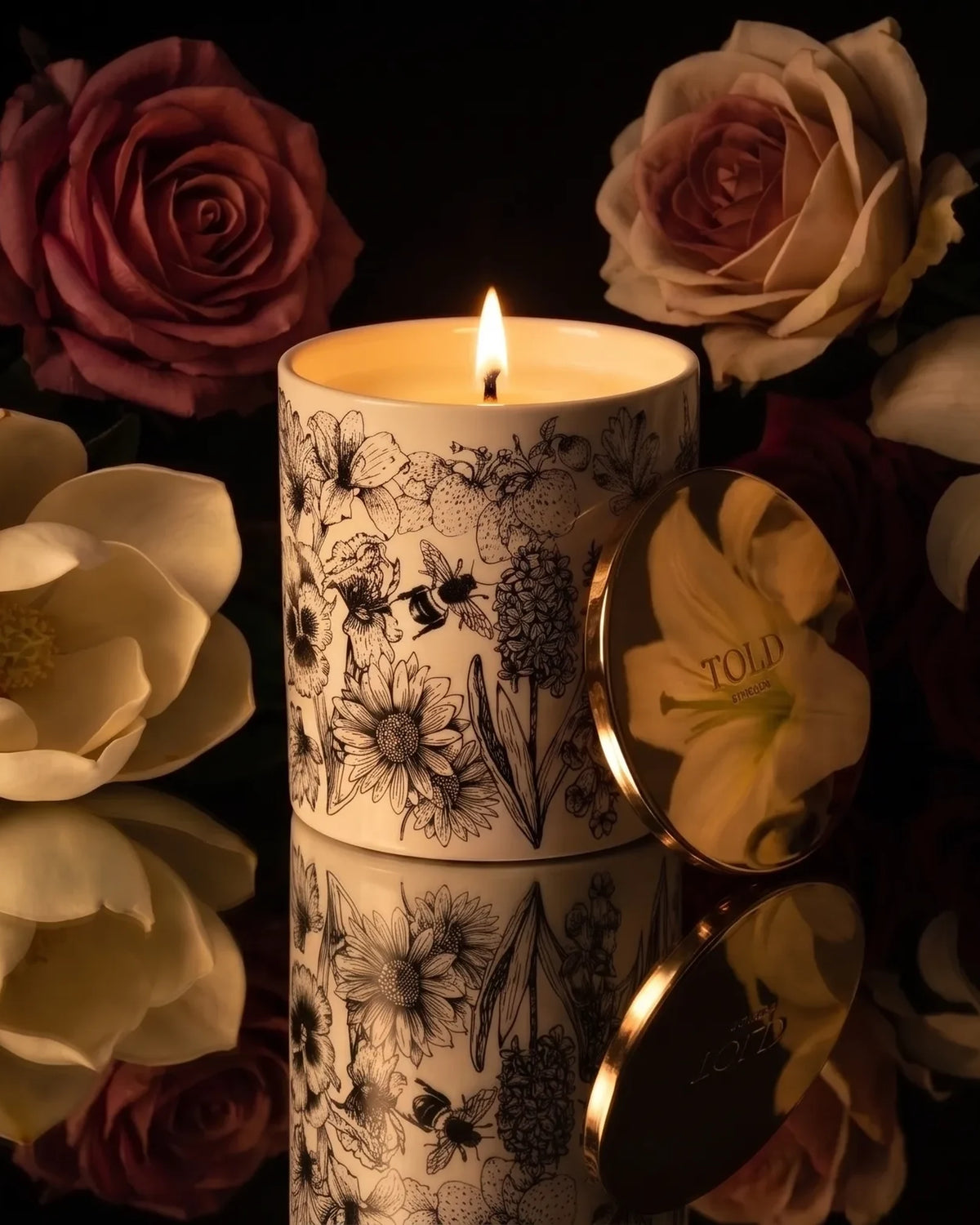 Una candela Told London accesa in porcellana illustrata, il coperchio dorato appoggiato accanto, tra rose, magnolie e gigli su una superficie a specchio scura.
