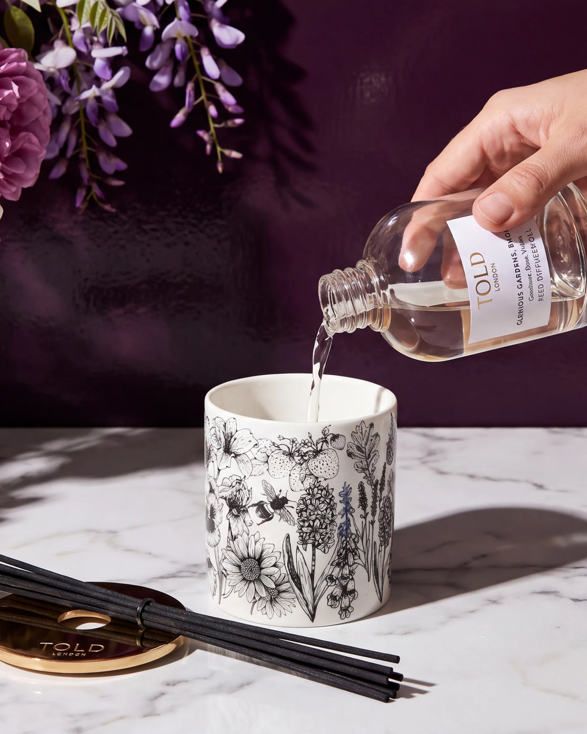 Olio per diffusore versato da un flacone di ricarica Told London in un vaso in porcellana illustrata su marmo bianco, con accanto il coperchio dorato e i bastoncini neri.