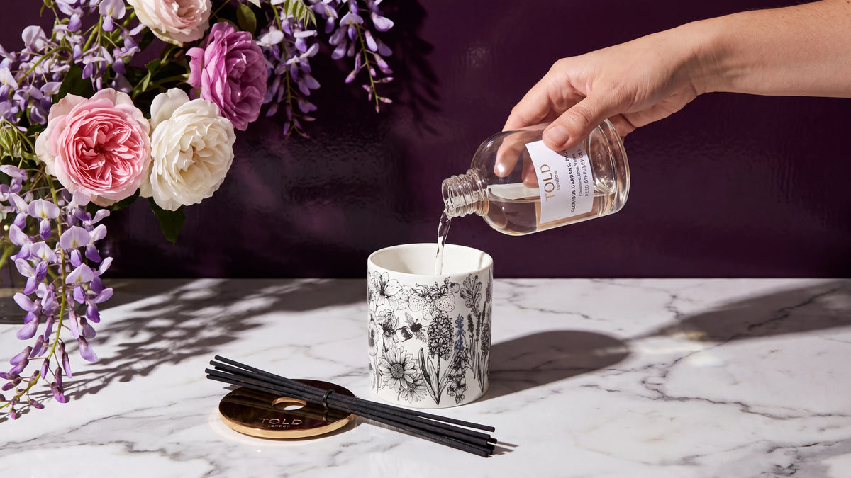 A hand pours reed diffuser oil from a Told London glass refill bottle into an illustrated porcelain diffuser vessel on white marble, with the gold lid and black reeds beside it and wisteria and garden roses behind.