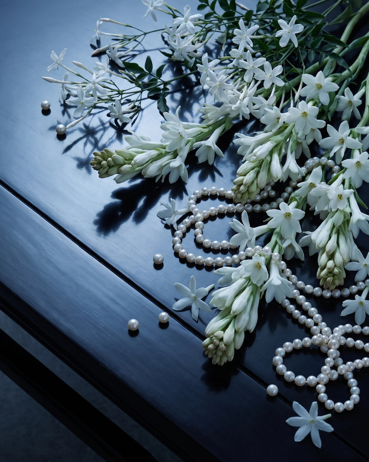 Sprays of white jasmine and strands of pearls on dark polished wood.