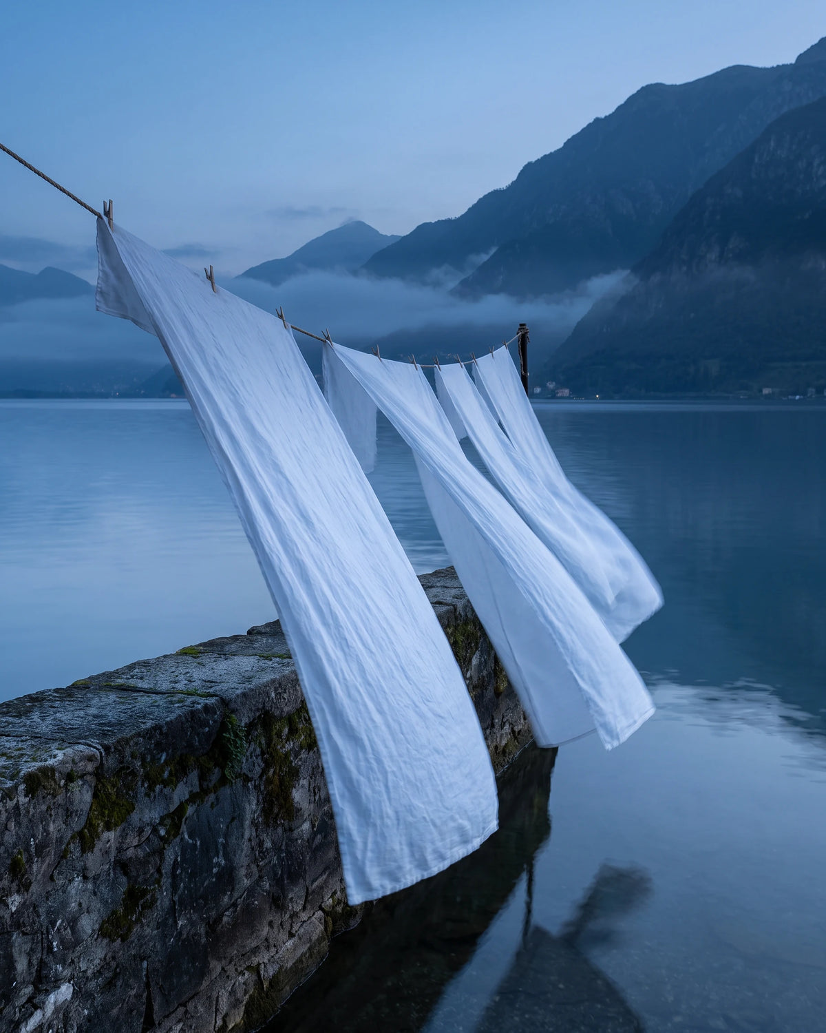 White linen hung on a line over a stone wall, lifting in the breeze, with mountains and mist across the lake.