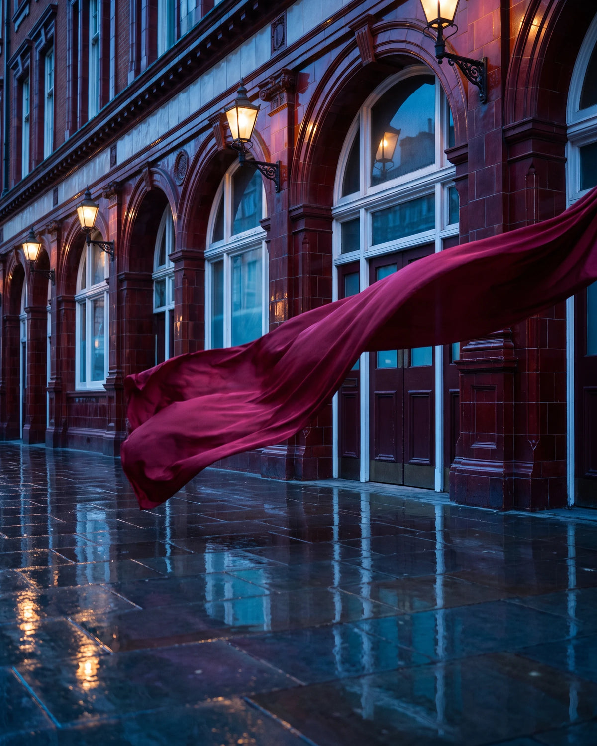 A rain-wet Victorian façade lit by lamps, oxblood silk caught mid-air in front of the arched windows.