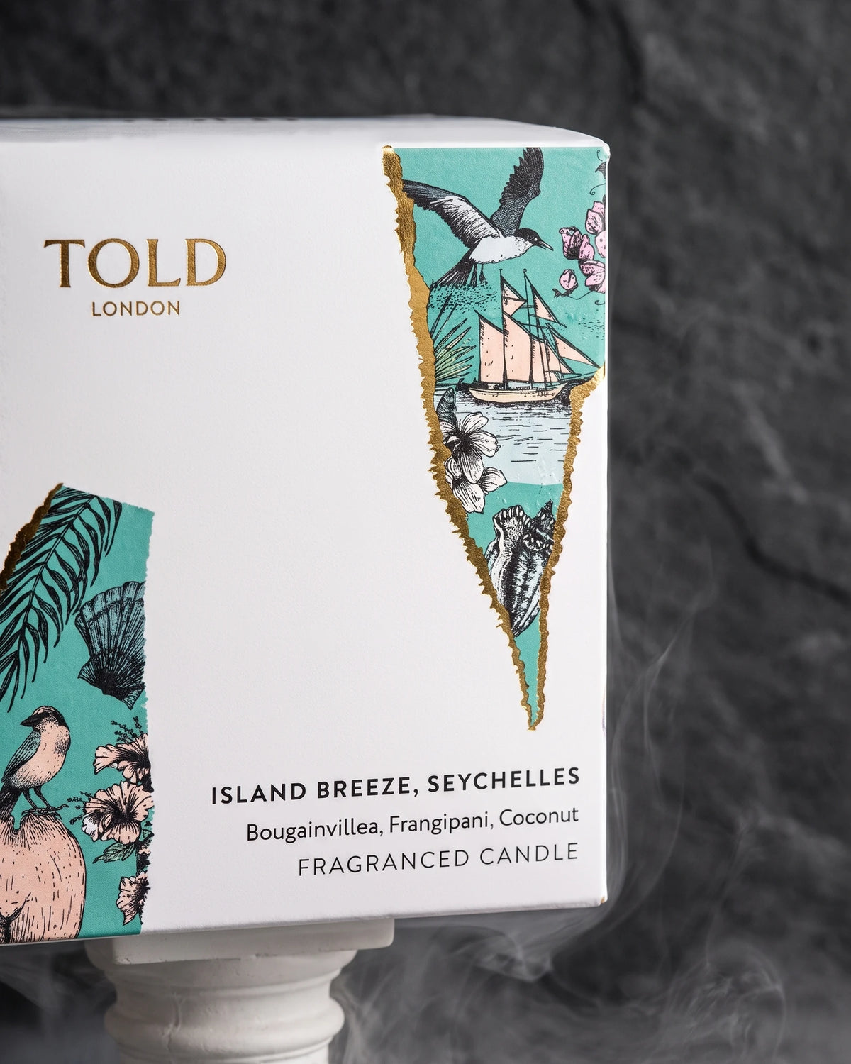 The Island Breeze candle box, white with torn strips of turquoise illustration edged in gold, on a white plinth.