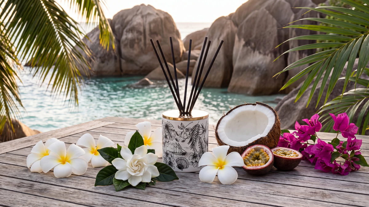 A Told London Island Breeze diffuser with black reeds on a weathered teak table above a Seychelles cove, with frangipani, a gardenia, a halved coconut, passion fruit and magenta bougainvillea, granite boulders and turquoise water behind.