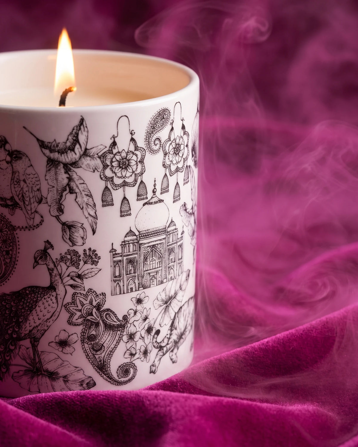 A lit Night Blooms candle in its illustrated porcelain vessel on fuchsia velvet, with a drift of pink smoke.