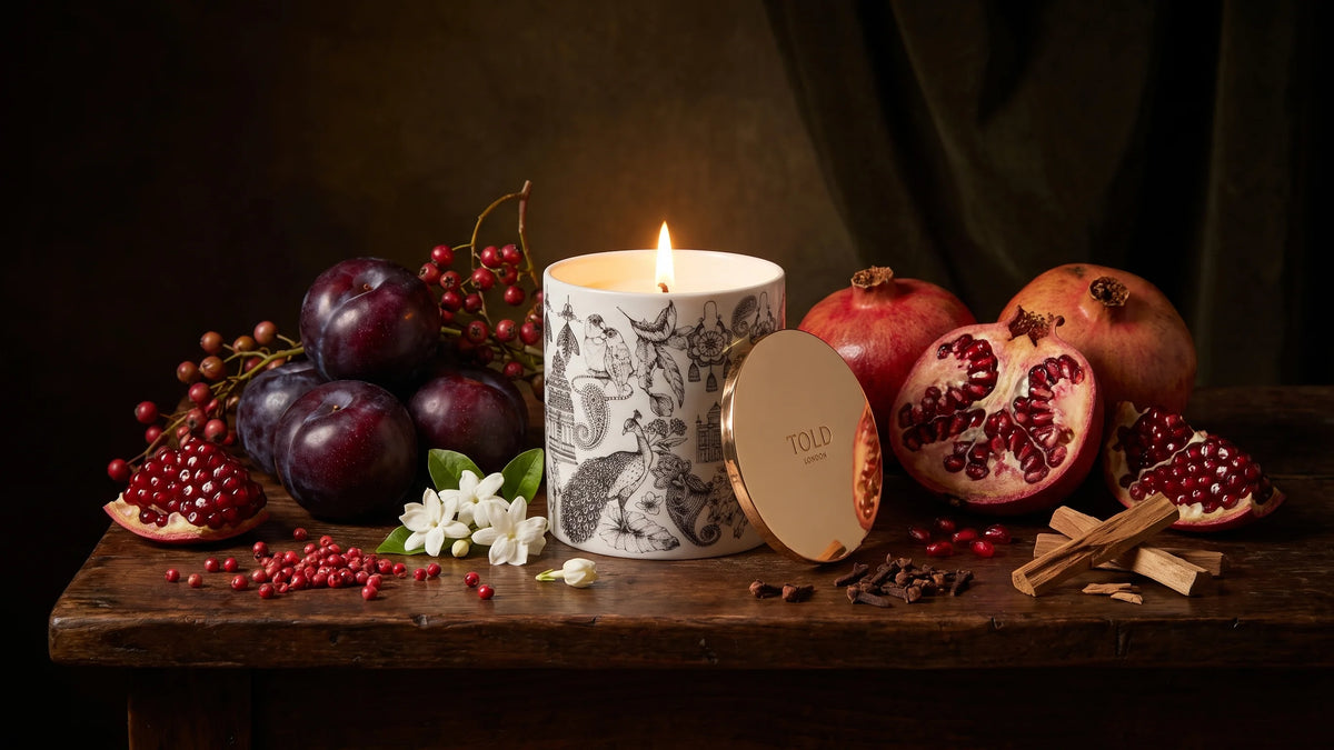 A lit Night Blooms, India candle on a dark wooden table with plums, pomegranates, jasmine, pink peppercorns, cloves and sandalwood.