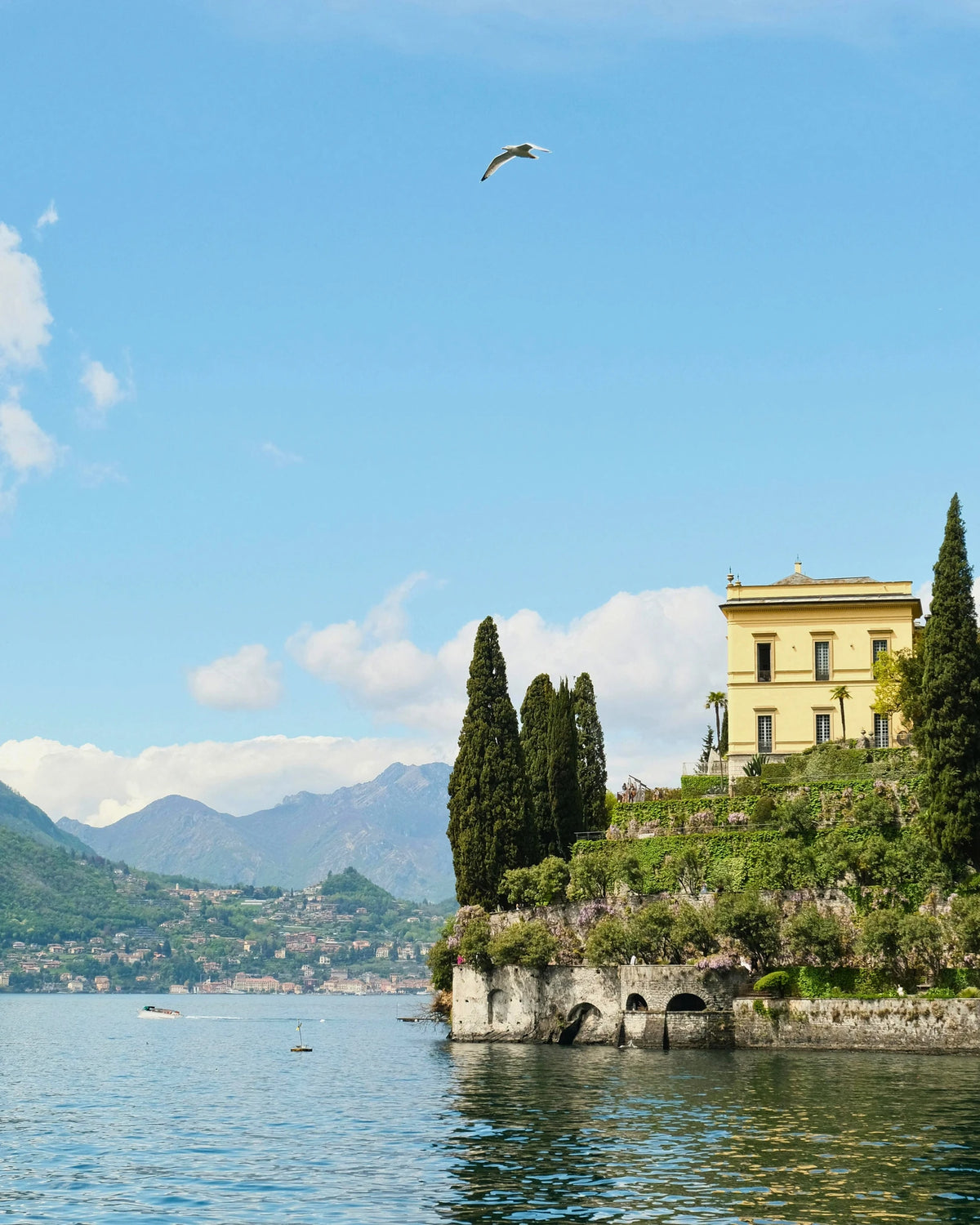 A yellow villa among cypress trees on a terraced point above Lake Como, with mountains beyond.